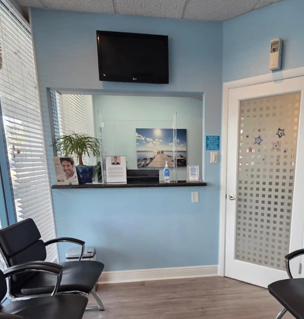 Dental office waiting area with black chairs, reception window, and a mounted TV on a light blue wall.