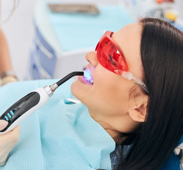 Dentist using an intraoral laser device with a blue light on a patient's teeth, patient wearing red protective glasses.