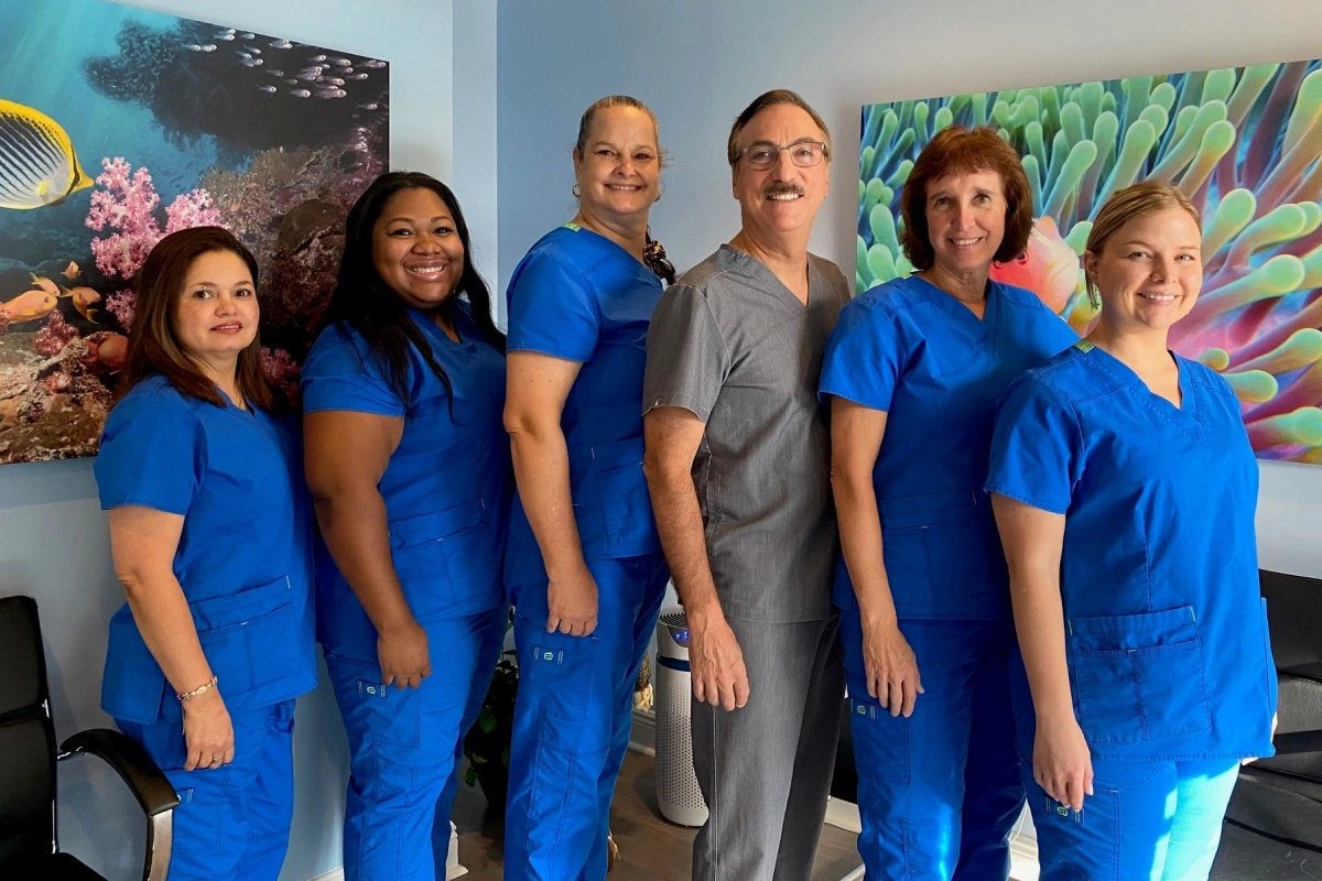 Dentist and five dental team members in blue and gray scrubs smiling together in front of ocean-themed wall art.