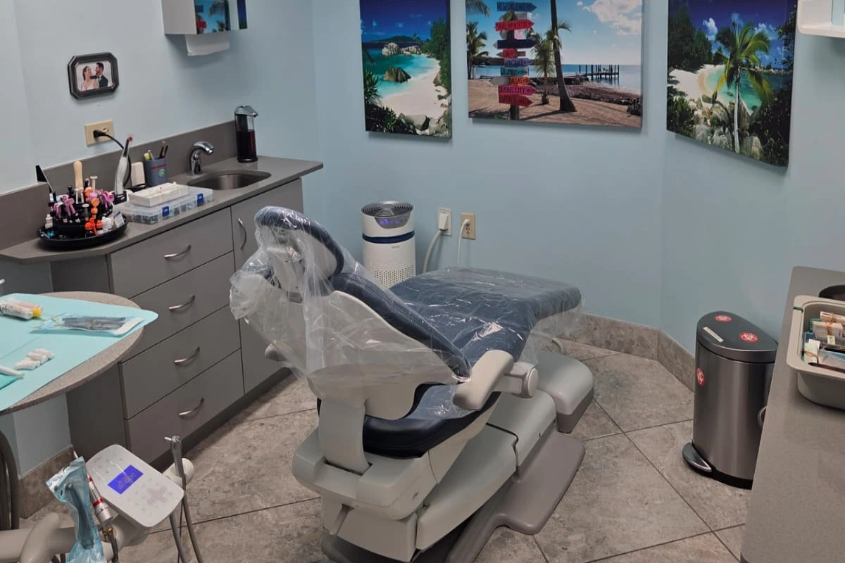 Dental treatment room with a covered patient chair, sink, and tropical beach-themed artwork on the walls.