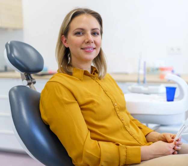 Smiling woman in a mustard-yellow blouse sitting in a dental chair, with dental equipment visible in the background.