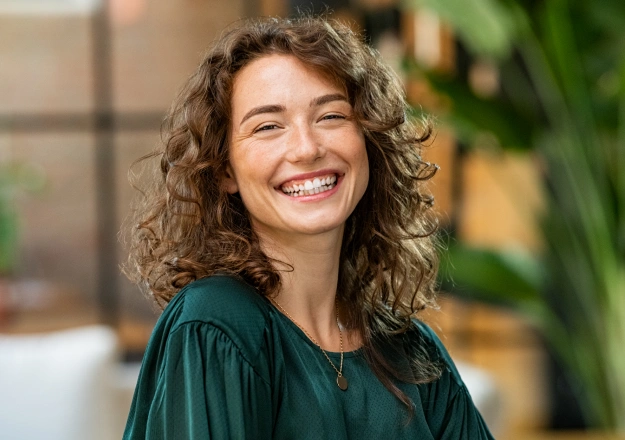 Smiling woman with curly hair wearing a green blouse, standing near a plant.