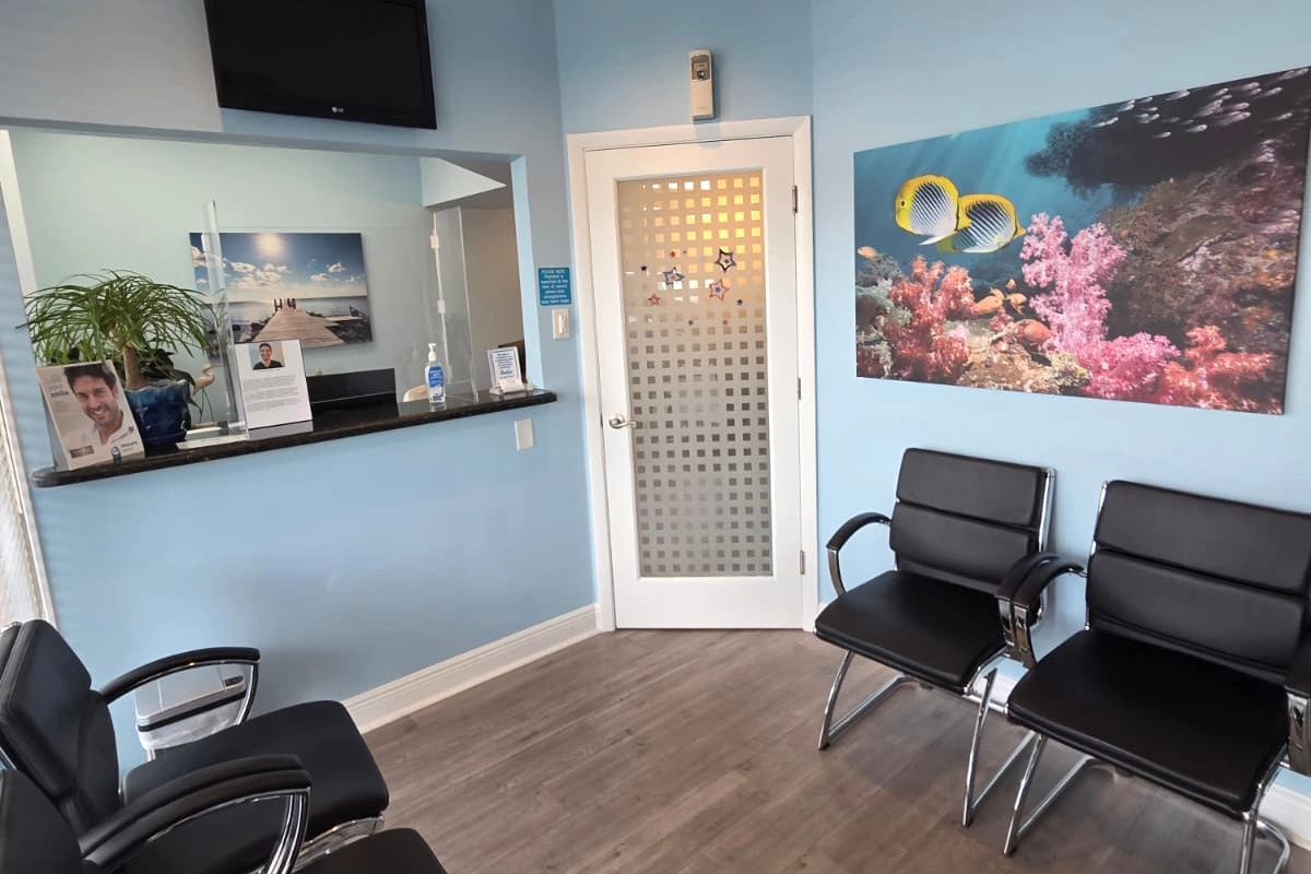 Dental office waiting room with black chairs, wood flooring, a reception window, and ocean-themed wall art.